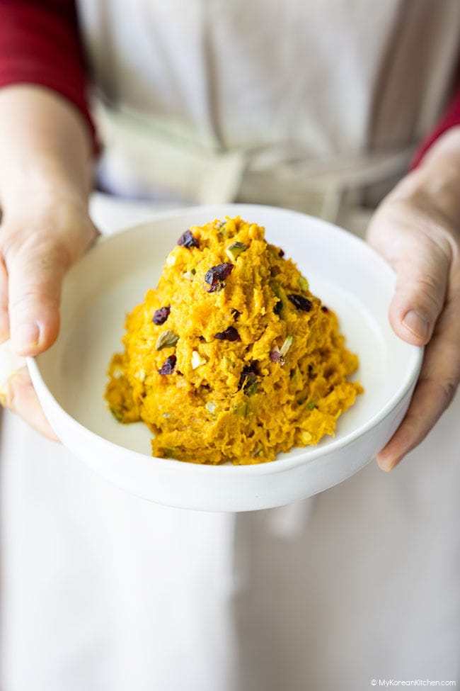 A bowl of Korean pumpkin salad topped with nuts and dried berries, held in both hands.
